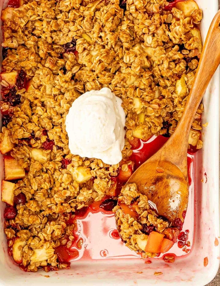 apple cranberry crisp in a casserole dish with a dollop of ice cream in the middle and a serving spoon inside.