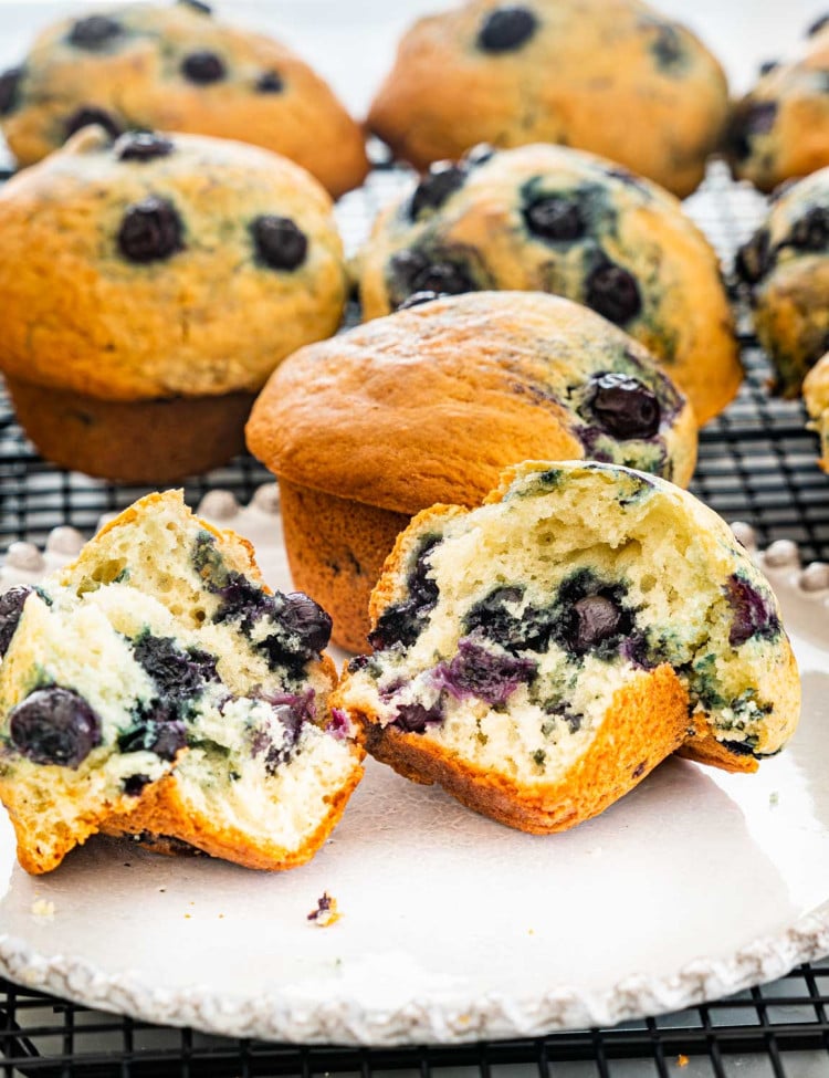 2 blueberry muffins on a plate, 1 cut in half with center exposed. More blueberry muffins in the background