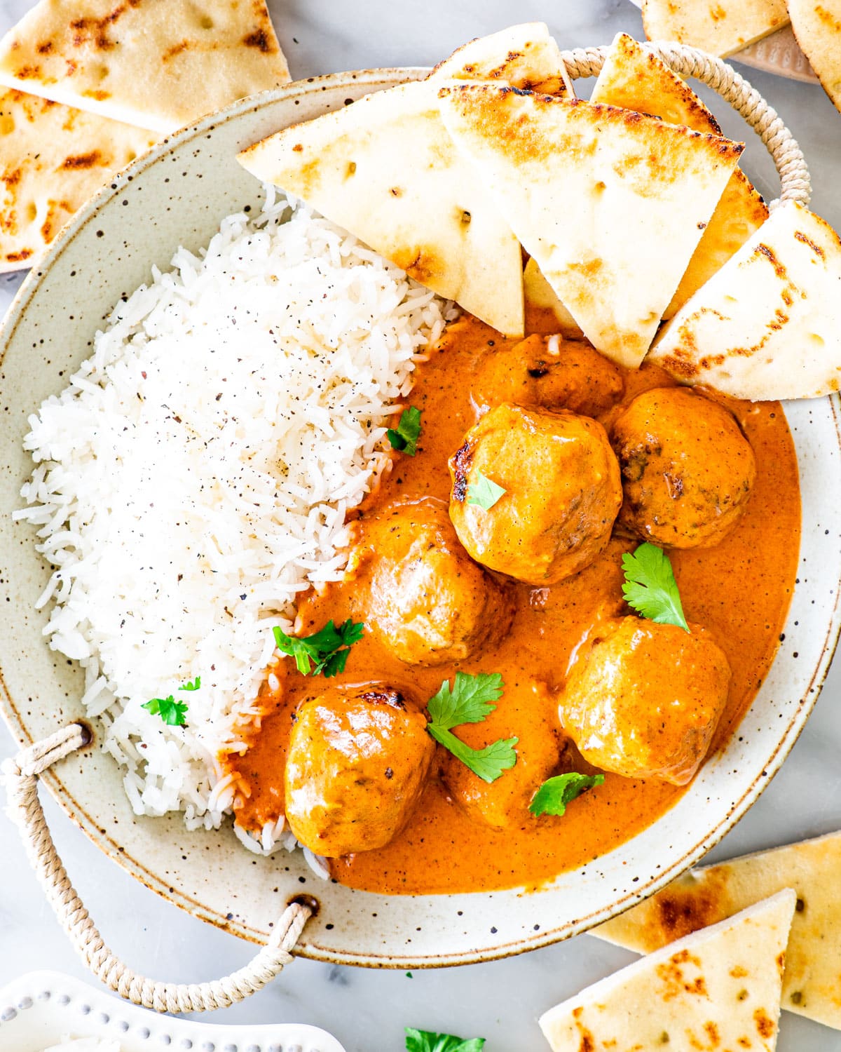 overhead shot of butter chicken meatballs with a side of rice in a bowl with naan slices and garnished with cilantro