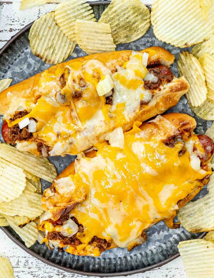 a couple freshly made chili dogs on a plate with potato chips.