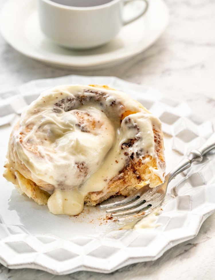 a cinnabon cinnamon roll on a white dessert plate with a cup of coffee in the background.