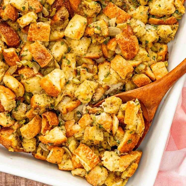 freshly baked bread stuffing in a white casserole dish with a wooden spoon inside.