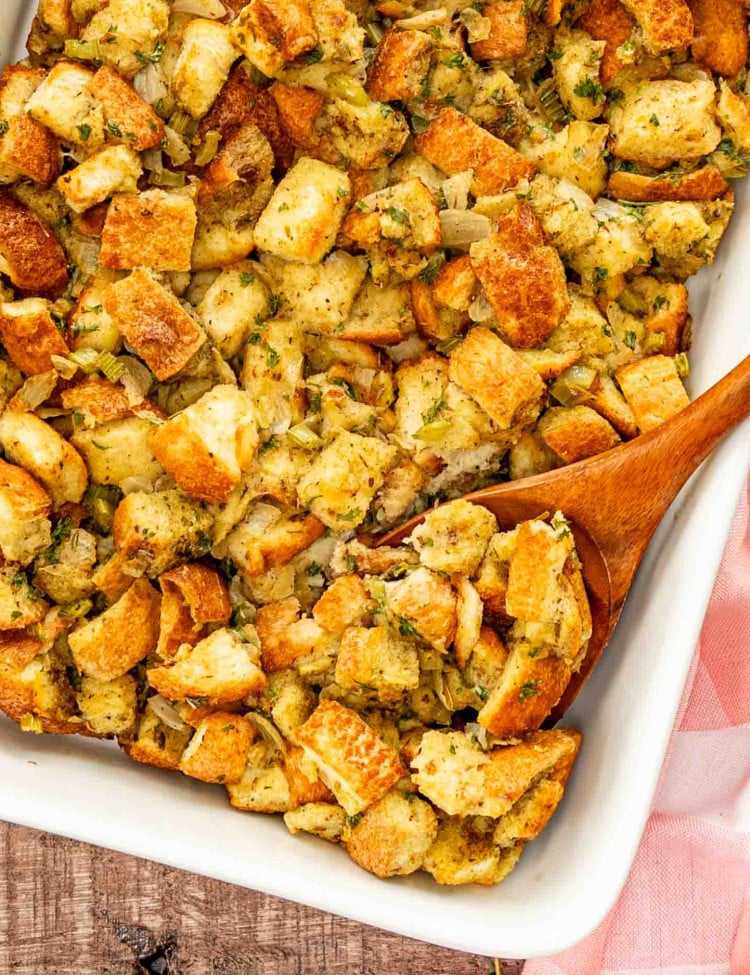 freshly baked bread stuffing in a white casserole dish with a wooden spoon inside.