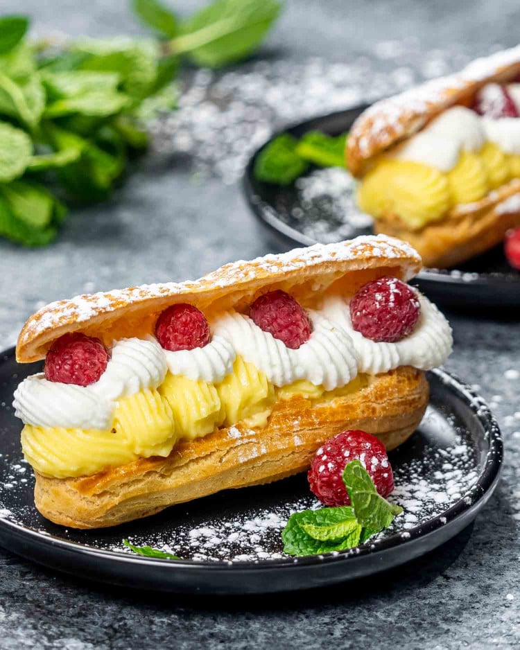 a gorgeous eclair filled with vanilla custard cream and topped with whipped cream and raspberries on a plate.