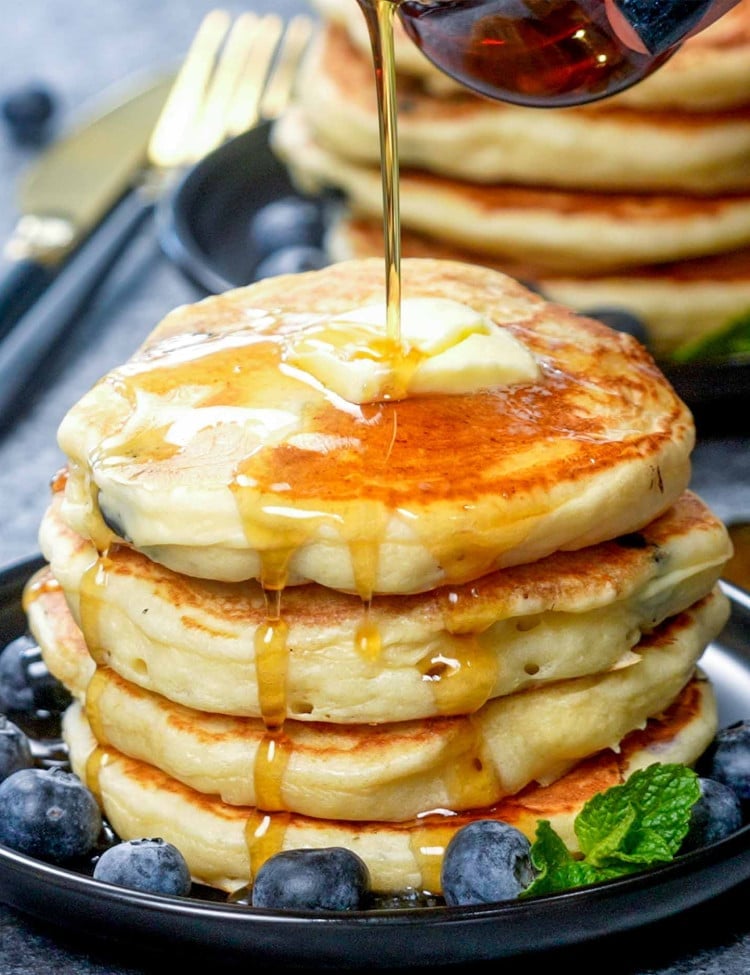 drizzling of maple syrup over a stack of lemon blueberry ricotta pancakes.