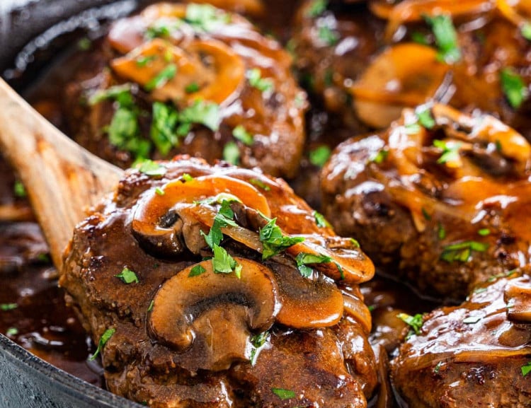 a wooden spoon lifting a salisbury steak out of the skillet